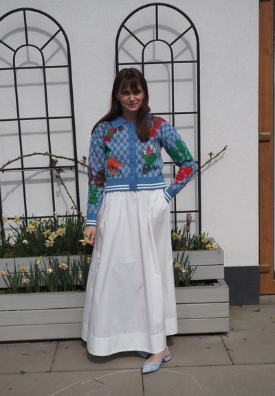 Mujer junto a una maceta con narcisos, lleva un cárdigan azul con estampado, una falda larga blanca y zapatos azul claro, delante de una pared blanca con enrejados metálicos.