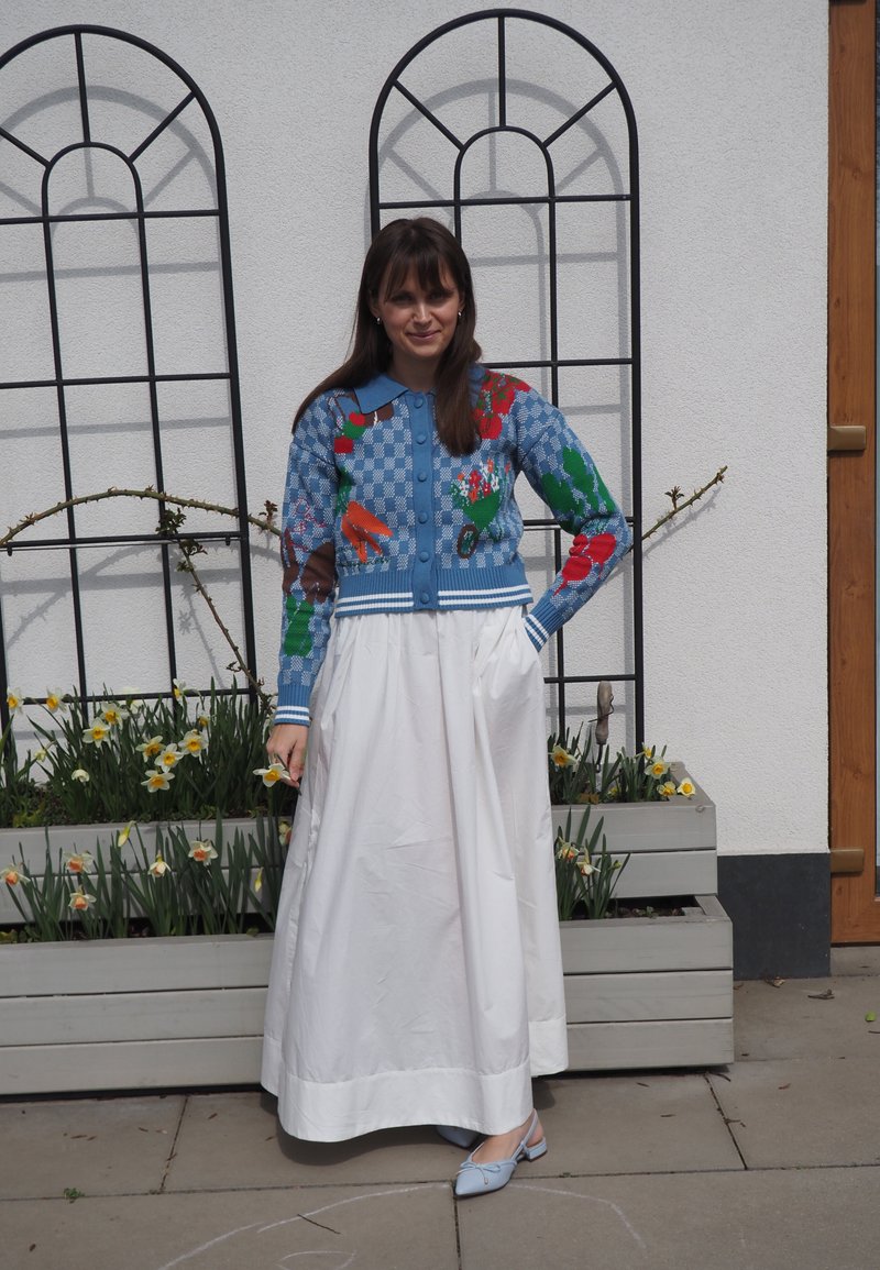 Femme debout à côté d'un bac à plantes avec des jonquilles, portant un cardigan bleu à motifs, une longue jupe blanche et des chaussures bleu clair, contre un mur blanc avec des treillis métalliques.