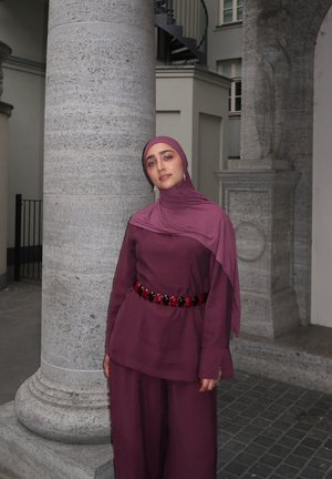 Mujer con un hijab burdeos y un conjunto a juego con un cinturón de cuentas, de pie junto a una gran columna de piedra en un entorno urbano al aire libre.
