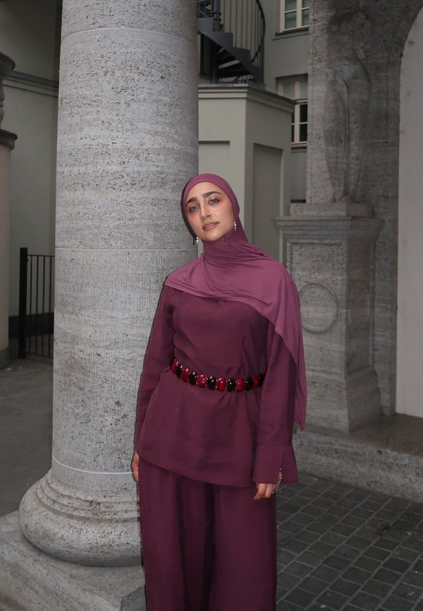 Kvinde iført en vinrød hijab og matchende outfit med et perlebesat bælte, stående ved en stor sten-søjle i et udendørs bymiljø.