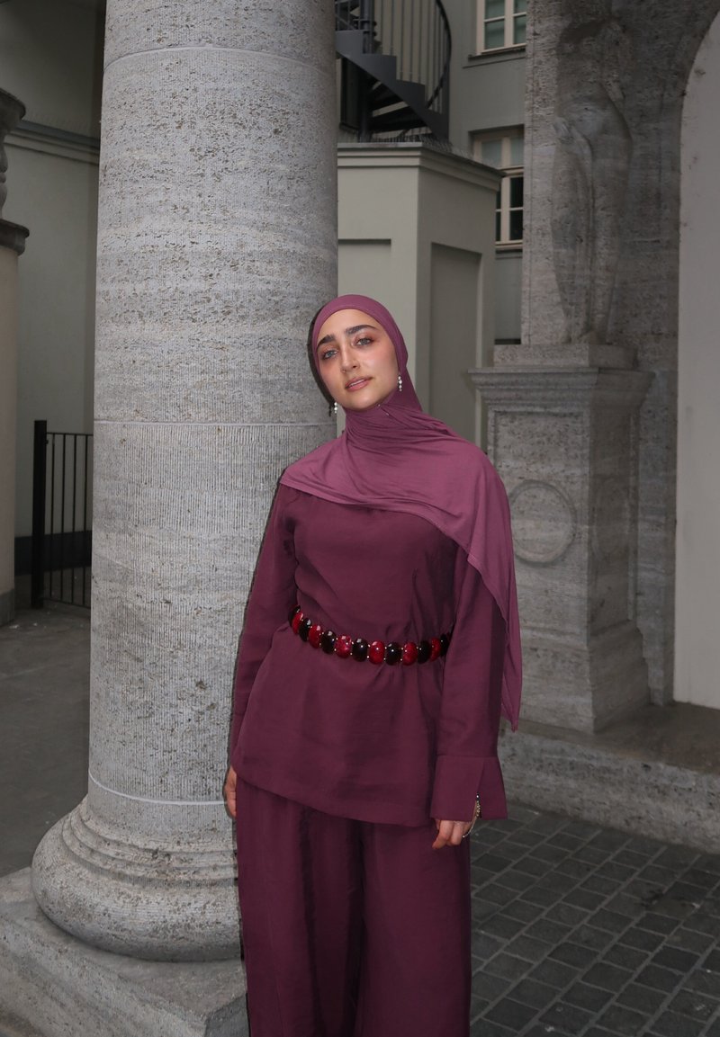 Frau mit einem kastanienbraunen Hijab und passendem Outfit mit einem Perlengürtel, stehend neben einer großen Steinsäule in einem städtischen Außenbereich.