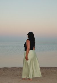 Top nero senza maniche e gonna lunga verde chiaro, in piedi su una spiaggia sabbiosa con un oceano calmo e un cielo pastello sullo sfondo.