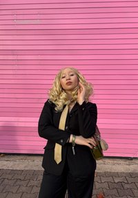 Juodas pritaikytas kostiumas su šviesiai ruda kaklaraiščiu. Ilgi, banguoti šviesūs plaukai. Žalia rankinė su pomponu. Fone matomas rožinės spalvos dryžuotas sienos paviršius.