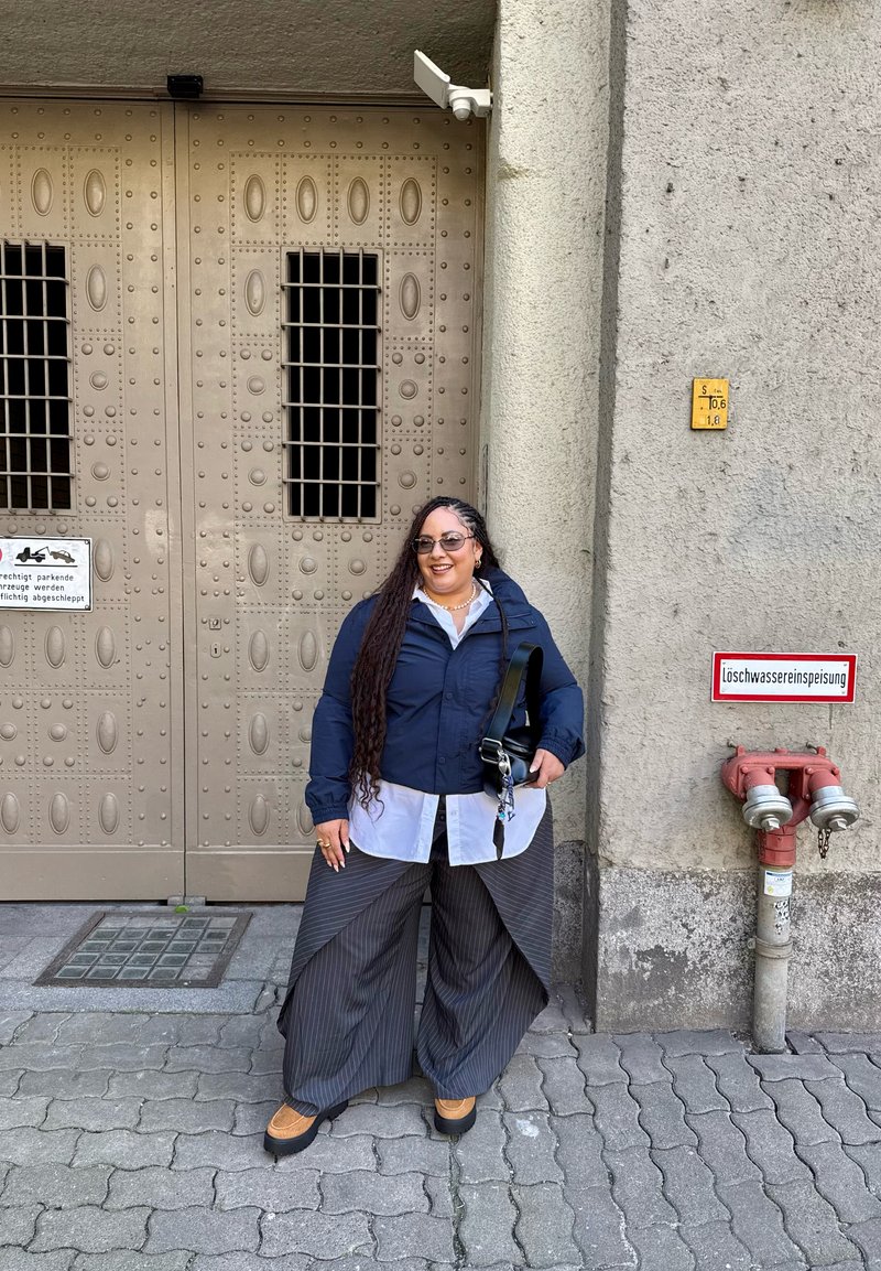Femme aux longs cheveux tressés se tient souriante devant une grande porte en métal et un mur en béton avec une bouche d'incendie et des panneaux d'avertissement.