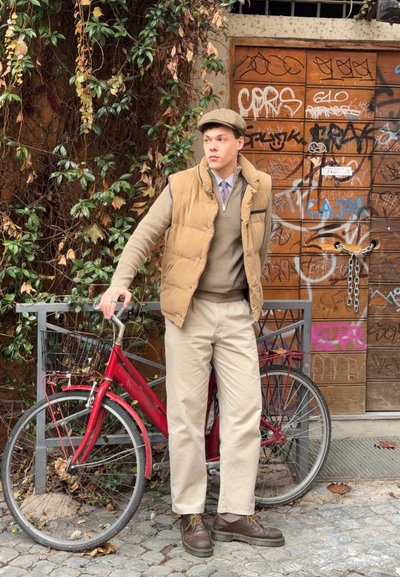 Chaleco acolchado beige, suéter marrón, pantalones kaki claros y zapatos oscuros. Bicicleta roja con una cesta delantera, apoyada contra una puerta de madera cubierta de grafitis.