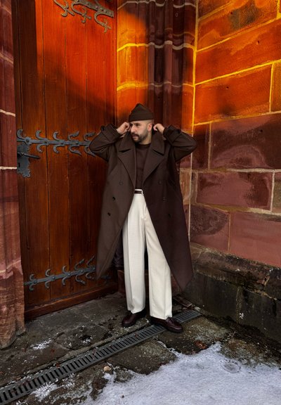 Hombre con abrigo marrón, gorro, pantalones blancos y zapatos oscuros ajustándose el sombrero, de pie junto a una gran puerta de madera y una pared de ladrillos rojos con nieve en el suelo.