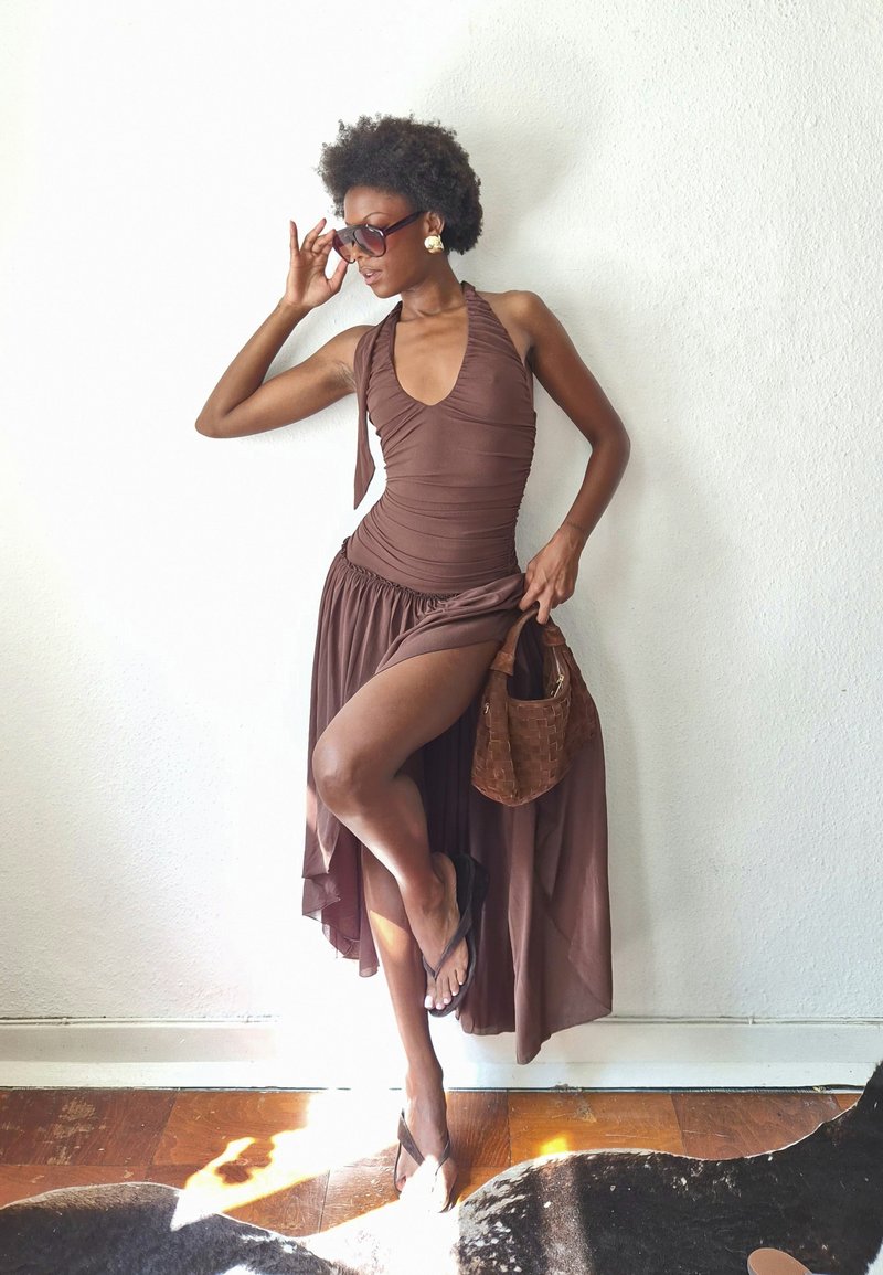 Femme en robe marron à col halter et sandales pose contre un mur blanc, tenant un sac à main marron tissé et ajustant de grandes lunettes de soleil.