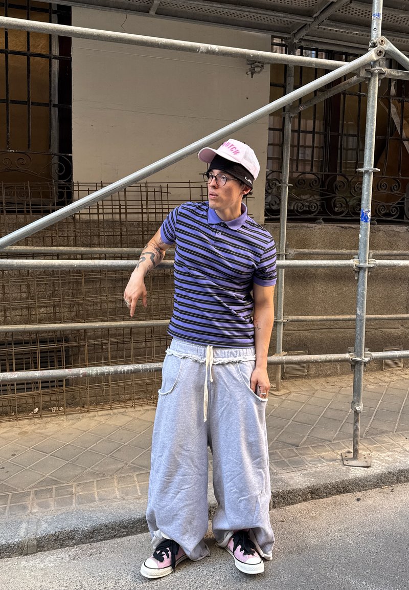 Polohemd mit lila und blauen Streifen, graue übergroße Sweatpants mit Kordelzug, schwarze und pinke Turnschuhe und eine pinke Baseballmütze mit schwarzen Akzenten.