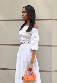 Weißes Off-Shoulder-Oberteil mit Puffärmeln, kombiniert mit einem weißen, gestuften Rock. Trägt eine kleine orangefarbene Handtasche mit geflochtenem Boden und Deckel.