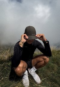 Musta pitkähihainen paita, tumma lippalakki, shortsit, valkoiset sneakerit. Taustalla ruoho ja pilvinen taivas. Henkilö kyykyssä kädet hatussa.