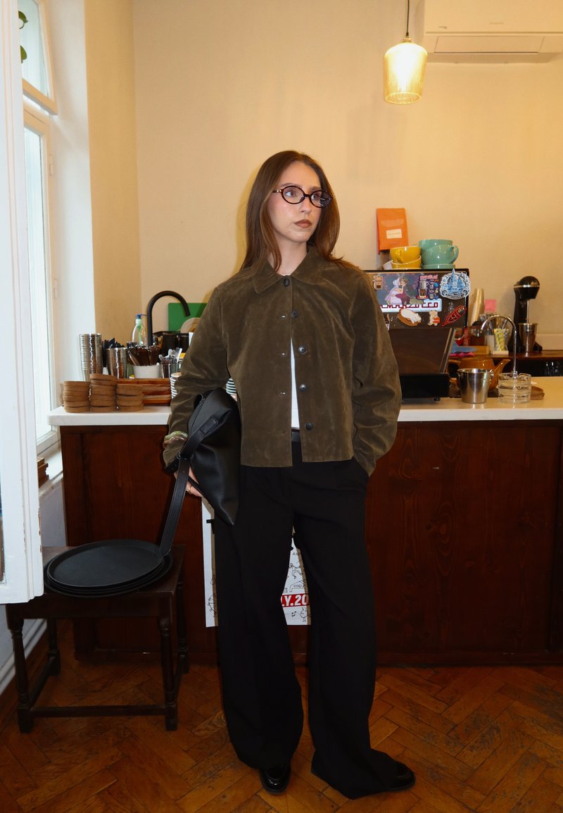 Young woman wearing glasses and a brown jacket holding a black bag stands near a café counter with cups and containers.