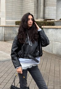 Femme aux longs cheveux noirs portant une veste en cuir noire et un jean gris, debout à l'extérieur tenant un sac à main noir près d'un bâtiment en pierre.
