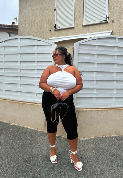 Top blanco con detalle de nudo, pantalones capri negros ajustados, tacones blancos con acentos de flores y un bolso negro pequeño. Cabello rizado estilizado con accesorios.