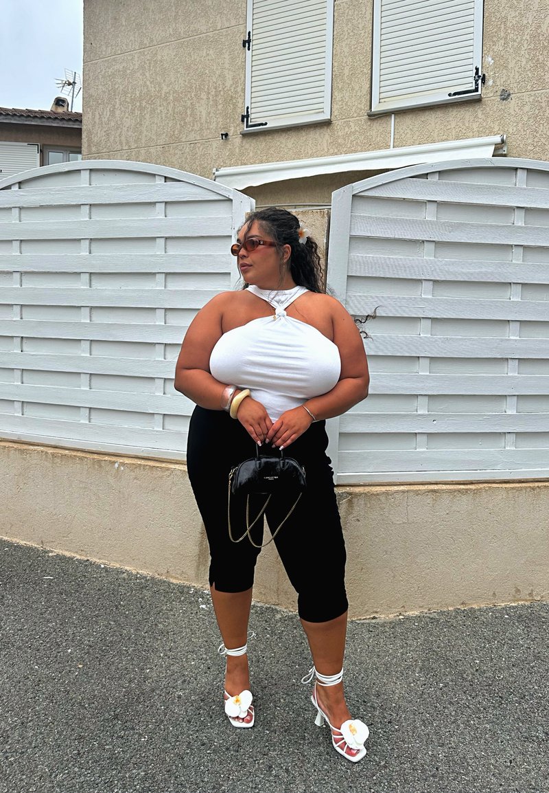 Haut halter blanc avec un détail en nœud, pantalon capri noir ajusté, talons blancs avec des accents floraux, et un petit sac à main noir. Cheveux bouclés coiffés avec des accessoires.