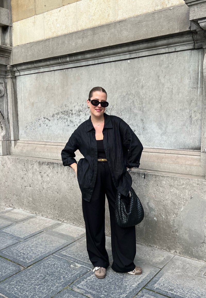 Femme portant une chemise noire surdimensionnée, un large pantalon noir, des lunettes de soleil, tenant un sac tissé noir, se tenant sur un pavé en pierre près d'un mur en béton gris.