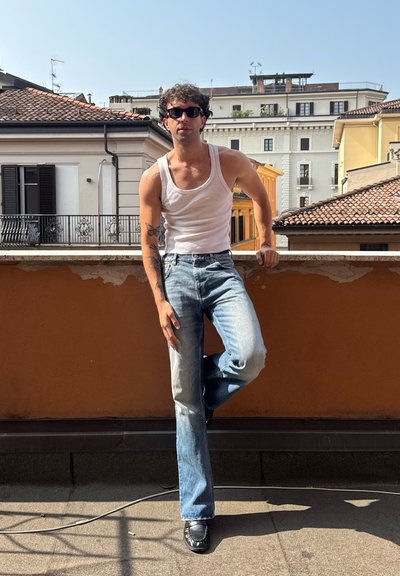 Camiseta blanca sin mangas, jeans azules desgastados y zapatos negros. Entorno al aire libre con un tejado de baldosas y edificios al fondo.