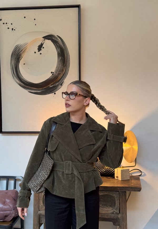 Femme aux cheveux tressés portant des lunettes, une veste ceinturée vert olive, et portant un sac à bandoulière à motifs, se tenant près d'une peinture abstraite ronde.
