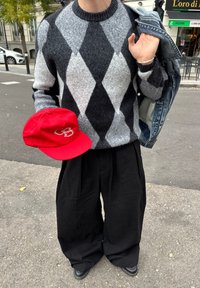 Personne portant un pull gris et noir à motifs losanges, un pantalon noir ample, tenant une casquette rouge dans une main et une veste en jean sur l'épaule.