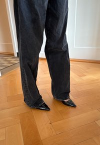 Chaussures à talons hauts noires à bout pointu, assorties à un jean denim foncé et ample. Sol en bois visible en arrière-plan.