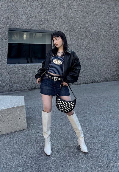 Chaqueta negra de gran tamaño, top corto a rayas, falda mini de mezclilla, botas blancas hasta la rodilla y un bolso negro con tachuelas. Texturas suaves y colores contrastantes.