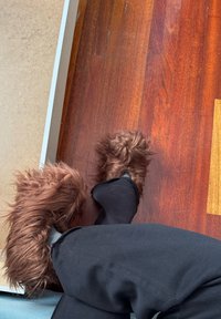 Bruine fuzzy slippers met een harige textuur, gedragen over zwarte broek, rustend op een houten vloer. De vorm van de slipper lijkt te groot.