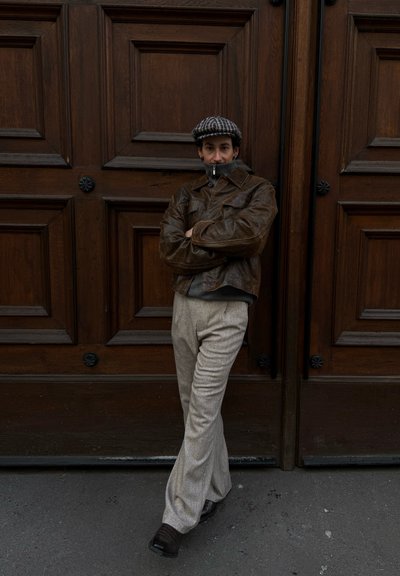 Persona con chaqueta de cuero marrón, pantalones grises y gorra estampada, apoyada contra una gran puerta doble de madera con los brazos cruzados.