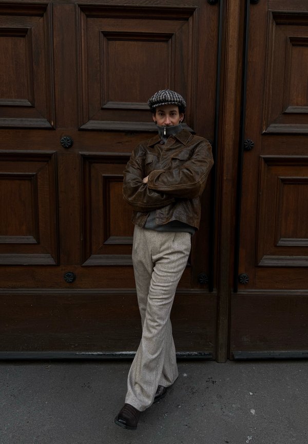 Osoba v hnědé kožené bundě, šedých kalhotách a vzorované čepici, opírající se o velké dřevěné dvoukřídlé dveře se založenýma rukama.