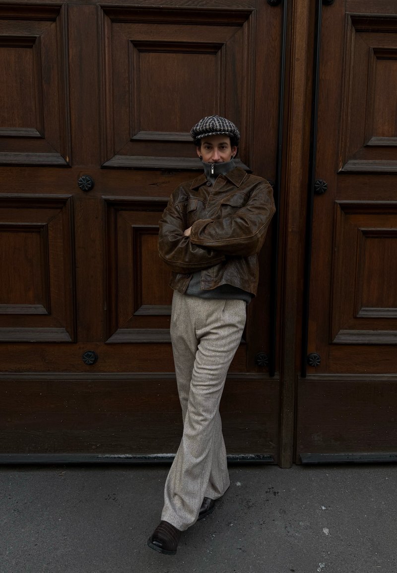 Osoba oblečená v hnedej koženej bunde, sivých nohaviciach a vzorovanej čiapke, opierajúca sa o veľké drevené dvojité dvere so založenými pažami.