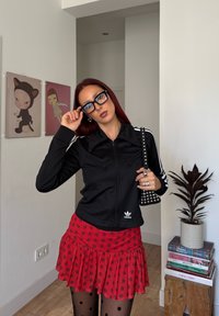 Kvinne med rødt hår iført svart Adidas-jakke, rød prikkete skjørt, svarte prikkete strømpebukser, som justerer brillene innendørs nær veggkunst og en plante.