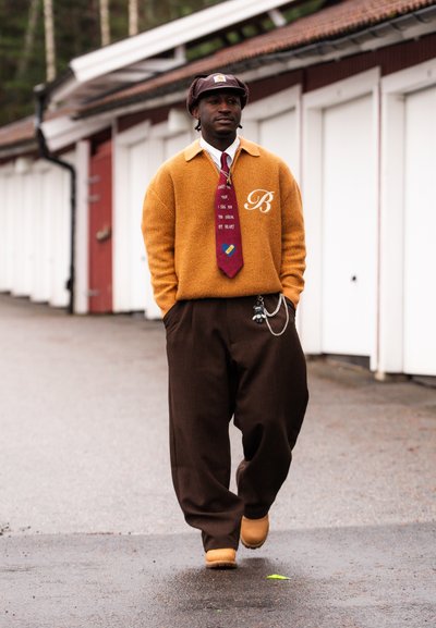 Hombre caminando al aire libre con suéter amarillo mostaza, corbata roja con gráfico de texto y pastillas, pantalones anchos marrones y sombrero marrón a juego, con las manos en los bolsillos.