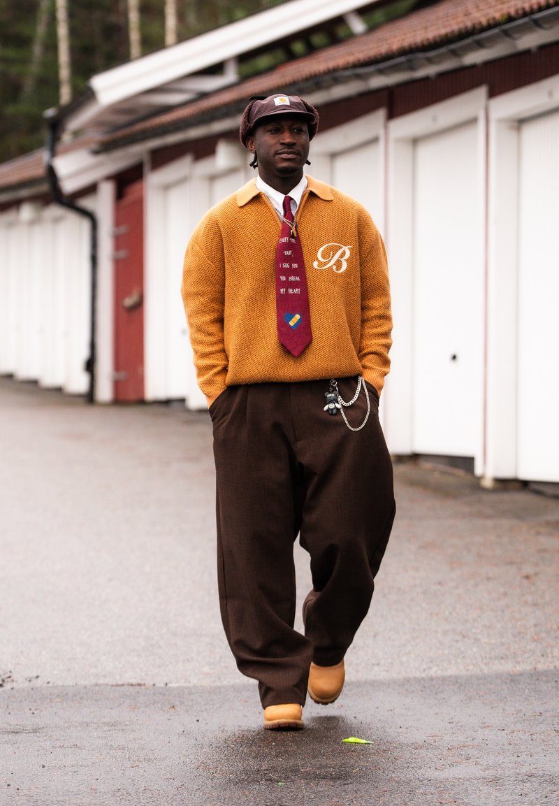 Muž kráčajúci vonku v horčicovo žltom svetri, s červenou kravatou s textom a grafikou tabliet, v hnedých širokých nohaviciach a zosúladenom hnedom klobúku, s rukami v vreckách.