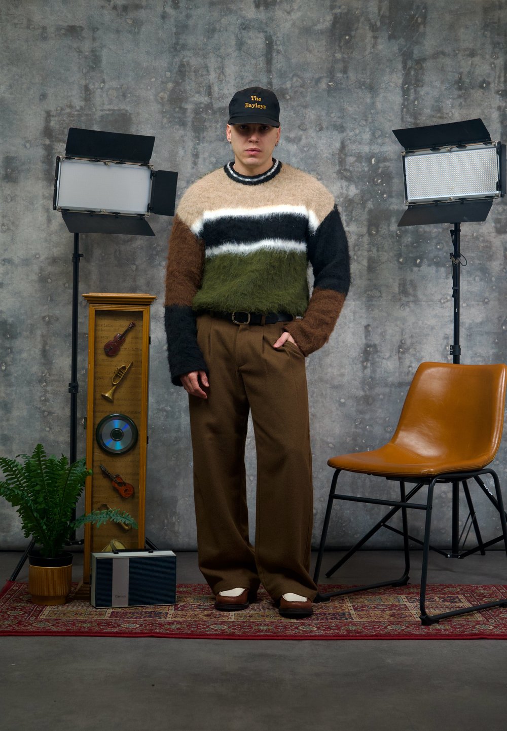 Pull en maille à rayures marron, vert, noir et crème ; pantalon large marron ; casquette noire. En arrière-plan, des éclairages de studio et des éléments décoratifs.
