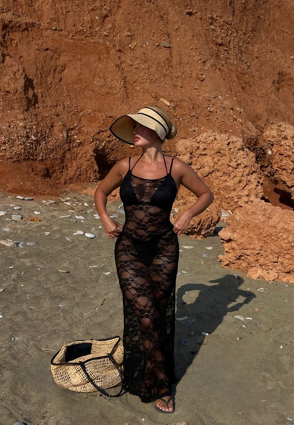Femme en robe noire en dentelle et grand chapeau de soleil se tient sur une plage rocheuse avec un sac tissé posé à côté d'elle par terre.
