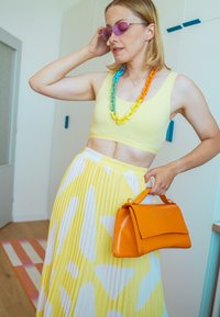 Gul crop top, plissert gul skjørt med hvite mønstre, oransje håndveske, og flerfarget kjedehalskjede. Lys bakgrunn og gulv.