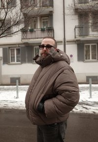 Άνδρας με μεγάλο καφέ μπουφάν τύπου puffer και γυαλιά ηλίου στέκεται σε χιονισμένο δρόμο μπροστά από κατοικημένο κτίριο τον χειμώνα.