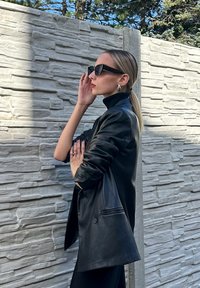 Blazer de pele preta com colarinho alto, tecido texturizado e corte ajustado. Usado com óculos de sol pretos e acessorizado com brincos de prata.