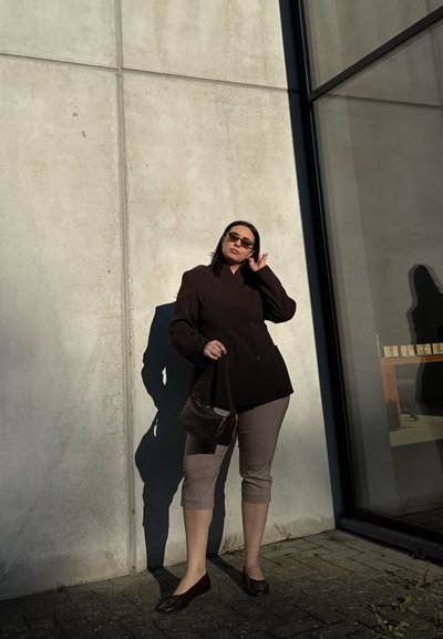 Mujer con blazer marrón, pantalones cortos a cuadros, gafas de sol y llevando un pequeño bolso, de pie contra una pared de hormigón bajo la luz del sol.