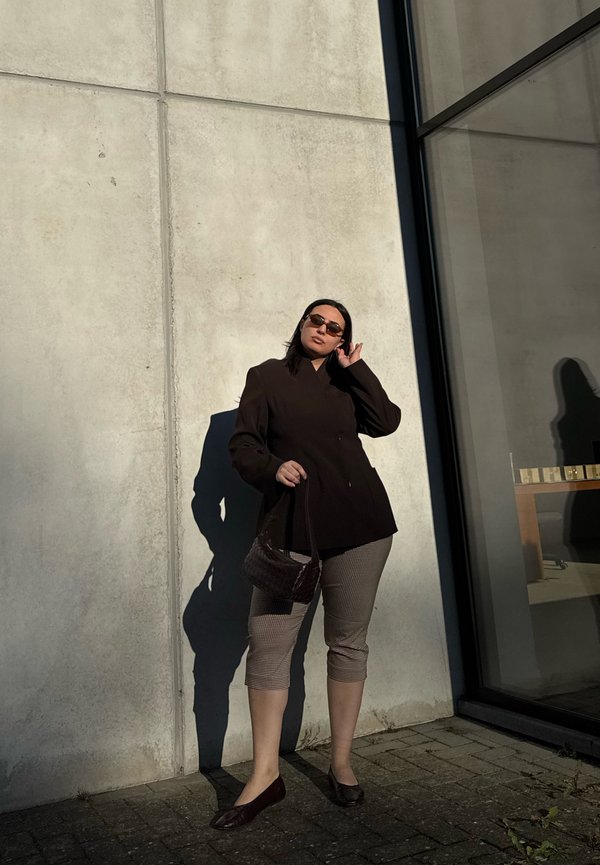 Žena s hnedým blejzrom, károvanými krátkymi nohavicami, slnečnými okuliarmi a nesúca malú kabelku, stojaca pri betónovej stene na slnku.