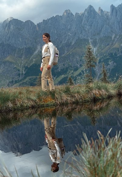 Escena al aire libre con una persona en pantalones beige y una camisa blanca de manga larga, de pie junto a un estanque que refleja montañas y vegetación.