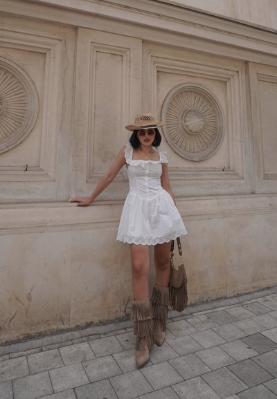 Vestido de algodón blanco con encaje, combinado con botas de flecos beige. Los accesorios incluyen un sombrero de paja y un bolso marrón con detalles de flecos.