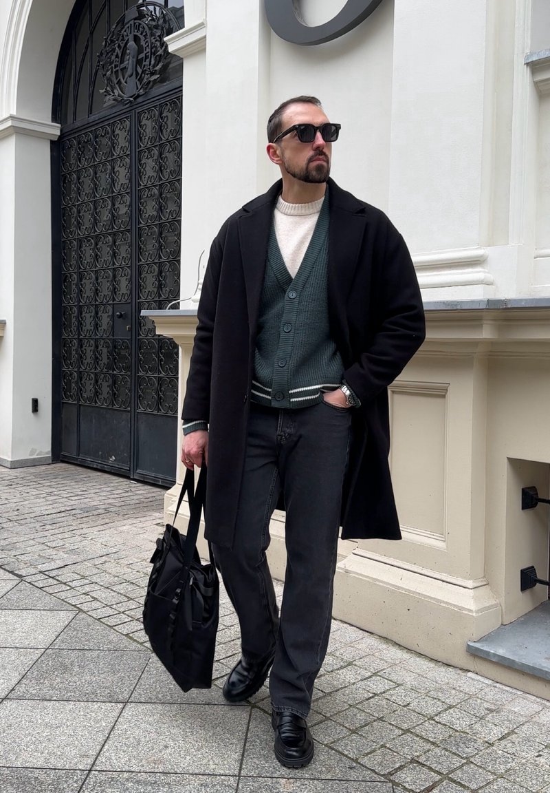 Homme portant un manteau noir par-dessus un cardigan vert et un pull blanc, jean noir et chaussures, portant un sac noir devant un mur de bâtiment.
