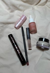 Kuusi meikkituotetta järjestettynä vaalealle kankaalle: Estee Lauderin huulituote (vaaleanpunainen), Lumenen nestemäinen poskipuna (pehmeä persikka), MACin eyeliner (musta), NARSin huulikiilto (läpinäkyvä) ja Bobbi Brownin voide (valkoinen).