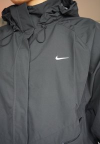 Chaqueta cortavientos negra con capucha ajustable mediante cordones y logotipo blanco de Nike en el pecho izquierdo. Material liso con diseño minimalista.