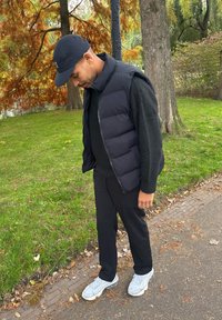 Doudoune sans manches noire portée sur un pull noir, associée à un pantalon noir et des baskets grises. Le décor comprend de la verdure et un feuillage d'automne.