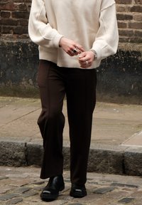 Pulover crem cu striații, pantaloni ajustați maro închis și bocanci cu talpă groasă negri. Ineluri din argint și un ceas pe mâini, fundal urban.