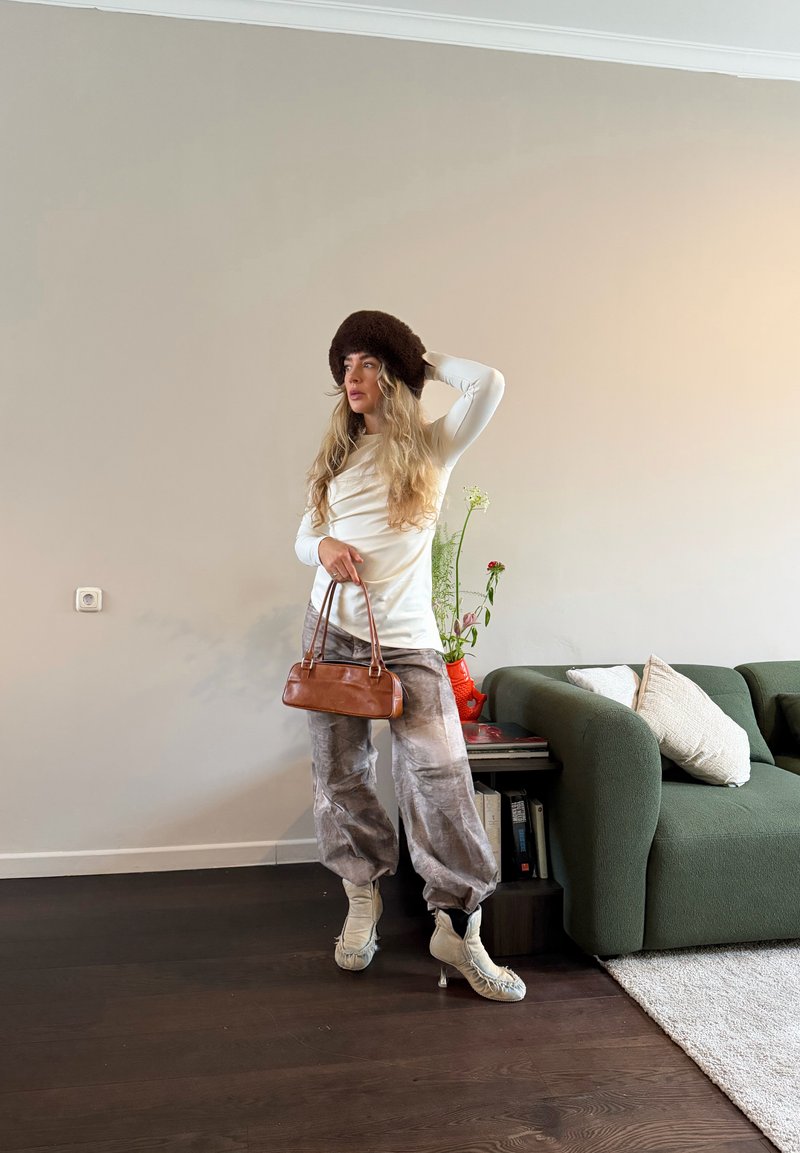 Femme en haut blanc, pantalon ample imprimé, chapeau en fourrure marron, talons beiges tenant un sac à main marron, debout près d'un canapé vert et d'une table d'appoint avec des fleurs.