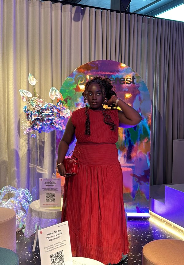 Femme en robe rouge plissée tenant une pochette rouge, debout devant un fond irisé Pinterest avec éclairage néon et fleurs décoratives.