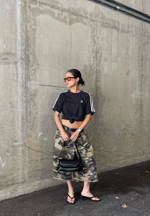 Donna che indossa una maglietta corta nera, una gonna midi camouflage, sandali neri con tacco e occhiali da sole arancioni, in piedi contro un muro di cemento tenendo una borsa nera.
