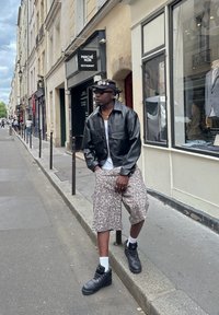 Homem vestido com uma jaqueta de couro preta, calções estampados e botas pretas posa numa rua de Paris em frente ao letreiro do restaurante "Marché Noir".