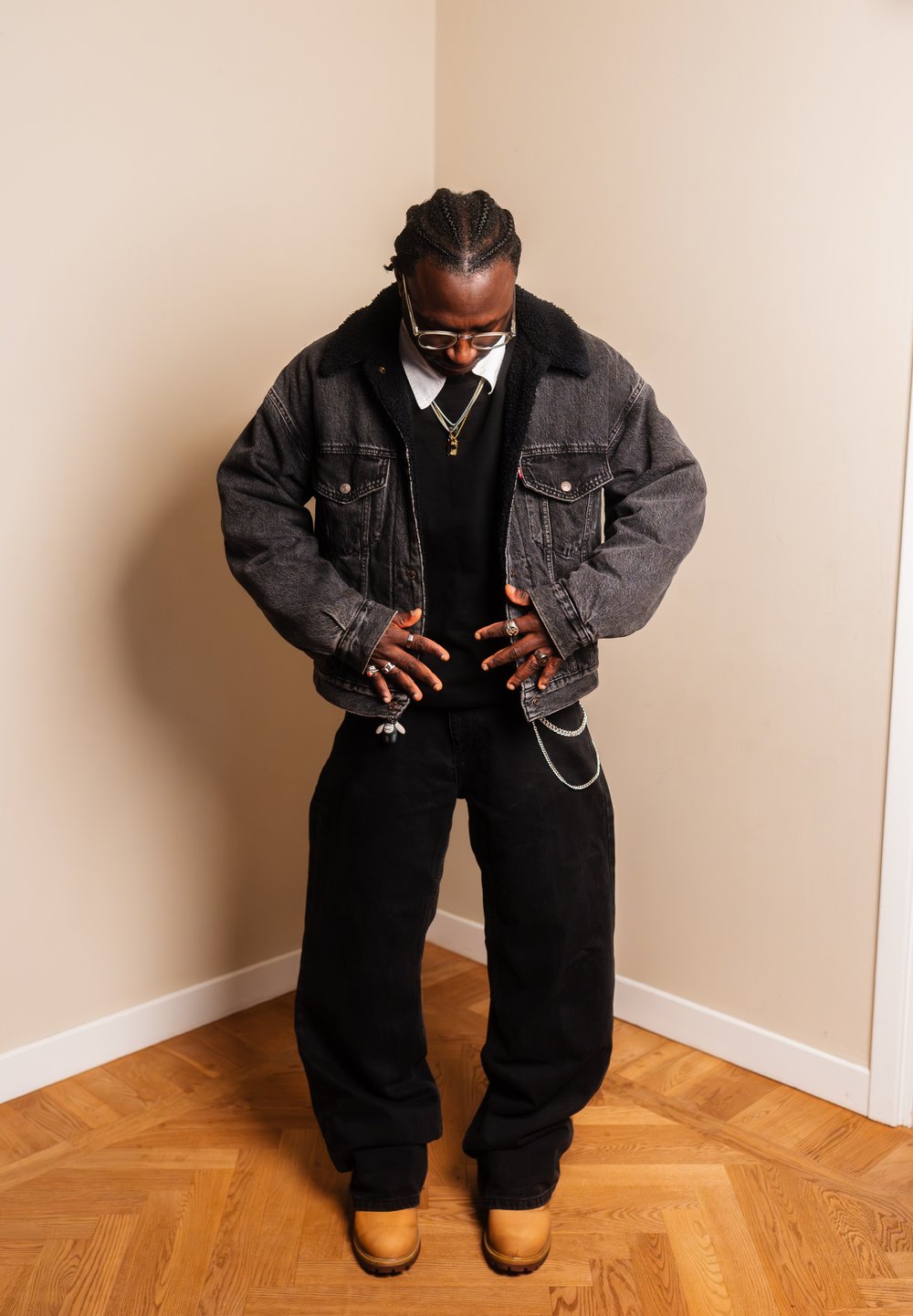 Homme aux cheveux tressés portant des lunettes, une veste en denim noire, un pantalon noir, des bottes couleur miel et plusieurs bagues, debout sur un sol en bois à l'intérieur.